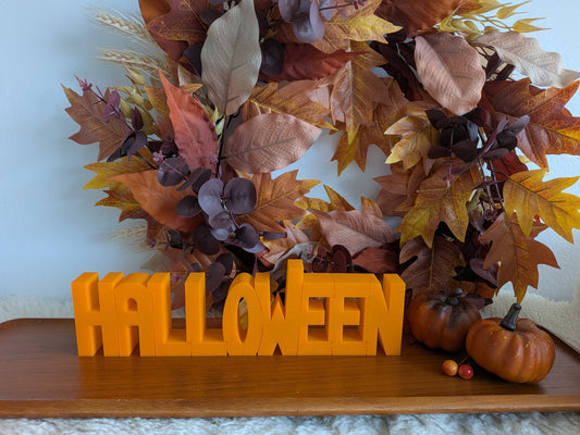 Orange 3D-printad dekorationsskylt med texten HALLOWEEN framför en höstkrans och pumpor